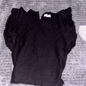 Svart volangblus från Holly & Whyte - Supersöt svart blus från Holly & Whyte med volangdetaljer på ärmarna och rund halsringning. Blusen har knapp i nacken för en snygg detalj och är tillverkad i ett mjukt tyg som känns skönt mot huden. Perfekt för dig som gillar stilrena plagg med en twist.