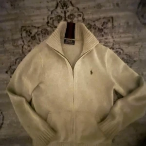 Beige stickad tröja Polo Ralph Lauren - Säljer en beige stickad tröja från Polo Ralph Lauren med halv dragkedja och ribbad krage. Tröjan har lång ärm, klassisk broderad logga på bröstet och snygga detaljer vid kragen. Perfekt för dig som gillar stilren och tidlös design.