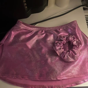 Glittrig rosa kortkjol från Mondor - Superfin glittrig rosa kortkjol från Mondor med matchande scrunchie. Kjolen har inbyggda trosor, konståkning/dans kjol! Vet inte vilken storlek men skulle säga att den passar folk med storlek 152-158! 