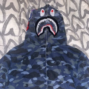BAPE blå camo Shark Hoodie - Säljer en ikonisk blå camo Shark Hoodie från BAPE med hel dragkedja, broderad haj på huvan och WGM-bokstäver i färg. Hoodien har känguruficka och är perfekt för dig som vill sticka ut med streetwear.