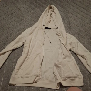 Beige hoodie från GGP Leisurewear - Säljer en beige hoodie från GGP Leisurewear i storlek M. Tröjan har dragkedja framtill, två fickor och justerbar huva med snörning. Tillverkad i mjuk bomull, perfekt för chill dagar. Klassisk passform och långärmad modell.