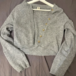 Grå croppad kofta med knappar - Supermjuk och croppad grå kofta från Gina Tricot med snygga träfärgade knappar framtill. Koftan är stickad, har lång ärm och en loose fit som ger en chill vibe. Perfekt att slänga över ett linne eller t-shirt för en avslappnad look.