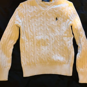 Vit kabelstickad tröja Polo Ralph Lauren - Kabelstickad vit tröja från Polo Ralph Lauren med klassisk rund halsringning och broderad marinblå logga på bröstet. Tröjan har långa ärmar och ribbade muddar för en snygg passform. Perfekt för lager-på-lager och stilrena outfits.