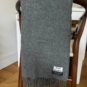 Grå halsduk från Acne Studios i ull - Stilren grå halsduk från Acne Studios i 100% virgin wool. Klassisk rektangulär form med fransar i ändarna och tydlig logotyp. Perfekt accessoar för att lyfta din vinterlook.