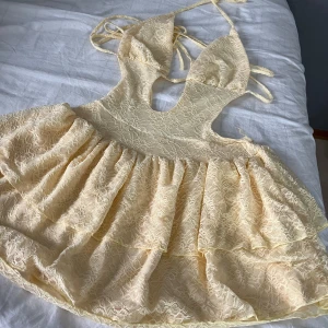 Ljusgul spetsklänning med volanger - Supersöt ljusgul kort klänning i spets med volanger och knyt i nacken. Klänningen har djup urringning, öppen rygg och är ärmlös. Perfekt för sommarens alla tillfällen när du vill sticka ut lite extra.