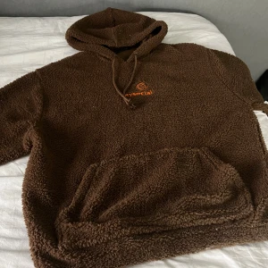 Brun fleecehoodie från Daysocial - Mysig brun hoodie från Daysocial i teddyfleece med broderad logga i orange, blått och grönt på bröstet. Tröjan har huva med snörning och stor magficka framtill. Perfekt för kalla dagar när du vill vara både bekväm och snygg.