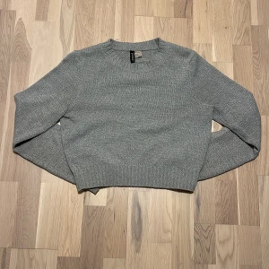 Grå croppad stickad tröja H&M Divided - Grå stickad tröja från H&M Divided i croppad modell. Tröjan har rund halsringning, ribbade muddar och lång ärm. Perfekt för lager-på-lager och chill dagar. Enkel och stilren design som funkar till det mesta.