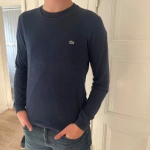 Mörkblå tröja från Lacoste - Säljer en stilren mörkblå tröja från Lacoste med klassisk rund halsringning och den ikoniska krokodilloggan på bröstet. Tröjan är långärmad och har en normal passform, perfekt för dig som gillar enkel och snygg stil.
