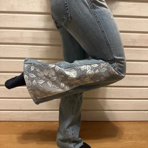 Blå bootcut jeans med spetsdetalj - Unika ljusblå jeans med bootcut-modell och breda spetsinslag i vitt med blommönster längs utsidan av benen. Jeansen har klassisk femficksdesign och är tillverkade i denim med snygg tvätt. Perfekt för dig som vill sticka ut med coola detaljer och vintagekänsla.