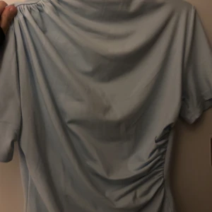 Ljusblå blus med rynk - Säljer en ljusblå blus med korta ärmar och snygga rynkade detaljer på sidan. Blusen har en enkel och modern design som passar perfekt för dig som gillar stilrena plagg med en twist. Materialet känns mjukt och skönt mot huden.