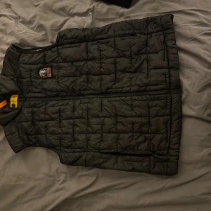 Svart quiltad dunväst från Parajumpers - Quiltad svart dunväst från Parajumpers med dragkedja framtill, ikonisk PJS-logga på bröstet och nacken samt orange detalj vid kragen. Västen har en sportig look och är fodrad i grått. Perfekt för lager-på-lager och streetstyle.
