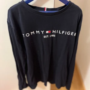 Blå långärmad tröja Tommy Hilfiger i storlek 152 - Svart långärmad tunnare tröja från Tommy Hilfiger med klassisk logga och texten 'EST. 1985' framtill. Tröjan har rund halsringning och är gjord i mjuk bomull, perfekt för en clean och stilren look.