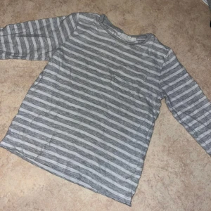 Randig långärmad tröja i bomull - Grå och vit randig långärmad tröja med rund hals och liten bröstficka. Tillverkad i mjuk bomull, perfekt för en chill och avslappnad stil. Passar lika bra till jeans som till mjukisbyxor.