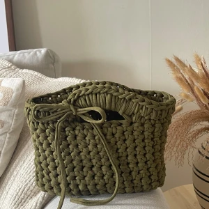 Olivgrön stickad handväska med knyt - Snygg handväska i olivgrönt, grovstickat material med charmig knytning framtill. Väskan har en mjuk, boxig form och är perfekt för dig som gillar unika accessoarer. Perfekt att bära till vardags för en cool och personlig stil.