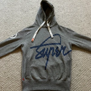 Superdry hoodie - Säljer en grå unik hoodie från Superdry med stort blått tryck både fram och bak. Tröjan har huva med vita snören. Skicket är som nytt har bara några gånger. Hör av er om ni har några frågor🤩🤩