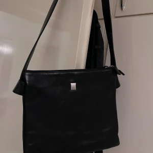 Svart axelväska från Gillian i skinn - Helt oanvänd. Snygg svart axelväska från Gillian i äkta skinn med stilren, rektangulär form. Väskan har flera fack med dragkedja både inuti och på baksidan, samt ett diskret metallmärke framtill. Perfekt för dig som gillar minimalistisk design och vill ha ordning på dina grejer.