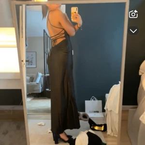 Svart långklänning med öppen rygg - Supersnygg svart långklänning i glansigt material med smala axelband och v-ringning. Klänningen har öppen rygg med snörning och sitter figurnära. Aldrig använt, Köpt ny för 600