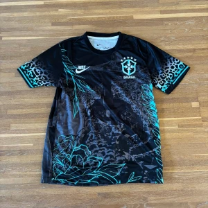 Nike Brasil svart fotbollströja M – unikt Rio-tryck & Dri-FIT (2) - Supersnygg svart Brasil-tröja från Nike med ikoniskt Rio de Janeiro-tryck och Kristusstatyn på framsidan. Detaljer i turkos, CBF-emblem på bröstet och leopardmönstrade ärmar som ger den en unik look.  ✔️ Lätt Dri-FIT-material som andas – perfekt både till träning och streetstyle ✔️ Korta ärmar, regular fit ✔️ I mycket fint skick  Passar dig som gillar fotboll, vintage streetwear eller bara vill ha en unik sporttröja. (Tröja nummer 2)