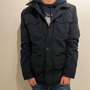 Mörkblå fieldjacka från J.Lindeberg - Snygg mörkblå fieldjacka från J.Lindeberg med fyra stora fickor framtill, knappar och hög krage. Jackan har diskret logga på ärmen och meshfoder på insidan. Perfekt för dig som vill ha en stilren och funktionell jacka med modern känsla. Två knappar har gått av men det är inget som syns eller märks🙌