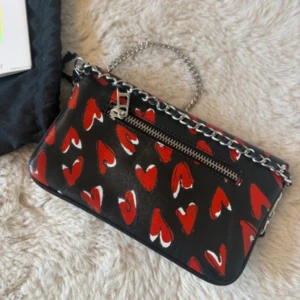Svart kuvertväska Zadig & Voltaire hjärtan - Kuvertväska i svart skinn från Zadig & Voltaire med röda hjärtan över hela väskan. Silverfärgad kedja som axelrem och dragkedja framtill. Insidan är röd och väskan har ett kuvertformat lock med magnetknäppning. Perfekt för dig som vill sticka ut. Endast använd vid 1 tillfälle! 