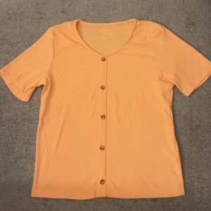 Persikofärgad ribbad topp från Primark - Säljer en persikofärgad ribbad topp från Primark med korta ärmar och u-ringning. Toppen har knappar framtill och är gjord i ett mjukt, stretchigt material som sitter skönt. Perfekt för dig som gillar en enkel men snygg stil. Säljer pågrund av att jag aldrig har använt den bara testat.