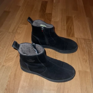Svarta fodrade boots med dragkedja Canada Snow - Svarta boots i mocka med mjukt, beige foder och dragkedja på sidan. Skorna har en platt sula med grovt mönster och en liten ögla baktill för enkel påtagning. Perfekta för kalla dagar när du vill hålla fötterna varma och bekväma.