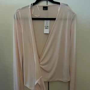 Ljusrosa omlottopp från Gina Tricot - Supersnygg ljusrosa omlottopp från Gina Tricot med långa ärmar och knyt i midjan. Tunn och mjuk i materialet, perfekt för lager-på-lager eller som den är. Djup v-ringning och stilren design som ger en fräsch look.