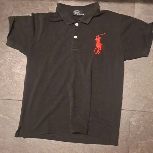 Svart piké från Polo Ralph Lauren - Svart pikétröja från Polo Ralph Lauren med röd broderad logga på bröstet och röda detaljer på ärmen. Klassisk krage, tre knappar framtill och custom fit. Perfekt för en clean och sportig look.