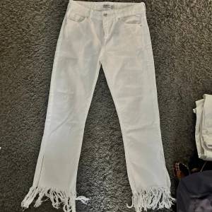 Vita jeans med fransar från Zara - Säljer ett par vita jeans från Zara Basic Z1975 Denim i storlek 36. Byxorna har raka ben med snygga fransar längst ner och klassisk femficksdesign. Materialet är denim och modellen är midwaist med dragkedja och knapp framtill. Liten i storlek