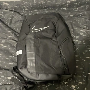 Svart ryggsäck  - Snygg svart Nike Elite ryggsäck med stor vit swoosh-logga och detaljer i silver. Ryggsäcken har flera fack, vadderade axelremmar med genomskinliga paneler och dragkedja. Perfekt för skolan eller gymmet, rymmer mycket och är riktigt stilren.