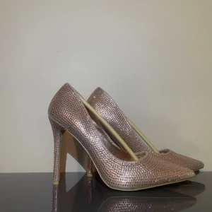 Glittriga champagne pumps - Säljer ett par glittriga pumps i champagne färg med spetsig tå och hög klack. Skorna är täckta av små glittrande stenar som ger en riktigt festlig look. Perfekta för dig som vill sticka ut och glänsa. Snyggt formade och med platt sula undertill.