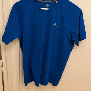 Blå Adidas t-shirt i bomull/polyester - Snygg blå t-shirt från Adidas med klassisk logga på bröstet. Tillverkad i en skön mix av 70% bomull och 30% polyester. T-shirten har rund halsringning och korta ärmar, perfekt för träning eller chill dagar. Storlek S.