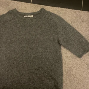 Grå stickad tröja från Zara - Liten i storleken så är närmare S! Mysig grå stickad tröja från Zara med rund halsringning och halvkort ärm. Perfekt för lager-på-lager och enkel att matcha med jeans eller kjol. Tröjan har en klassisk och stilren look som funkar året runt.