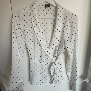 Prickig omlottblus från Gina Tricot - Säljer en vit omlottblus med svarta prickar från Gina Tricot. Blusen har långa ärmar, knyt i midjan och en snygg v-ringning. Perfekt för dig som gillar klassiska mönster med en modern twist.