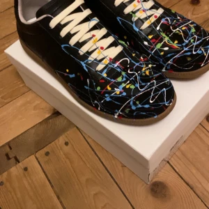 Svarta sneakers med färgstänk - Unika svarta sneakers med coolt färgstänksmönster i blått, vitt, rött, gult och grönt. Skorna har vita snören och en klassisk rund tå. Snygg kontrast mellan den svarta basen och de färgglada detaljerna. Perfekt för dig som vill sticka ut med din stil.