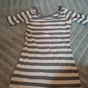 Randig kortklänning från Vero Moda - Snygg randig kortklänning från Vero Moda Basics med breda grå och vita ränder. Klänningen har rund halsringning och trekvartsärm. Tillverkad i mjuk bomull med lite stretch för skön passform. Perfekt för dig som gillar enkel och stilren design.