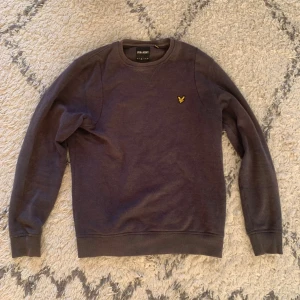 Mörklila sweatshirt från Lyle & Scott - En stilren mörklila sweatshirt från Lyle & Scott med rund hals och klassisk gul logga på bröstet. Tröjan har långa ärmar och ribbade muddar vid ärmslut och nederkant. Perfekt för en chill och avslappnad look.