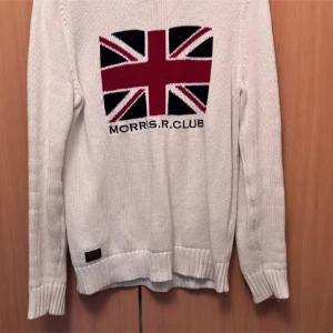 Vit stickad tröja Morris R Club S - Vit stickad tröja från Morris R Club med Union Jack-flagga och text framtill. Tröjan har ribbade muddar vid ärmslut och nederkant samt lång ärm. Perfekt för dig som gillar klassisk brittisk stil och vill sticka ut lite extra.