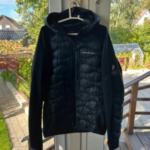 Svart pufferjacka Peak Performance L - Svart pufferjacka från Peak Performance i storlek L. Jackan har huva, dragkedja framtill och vadderade partier på bröst och ärmslut. Materialet är en mix av polyester och syntet, perfekt för kyliga dagar. Snygg och sportig look med diskret logga på bröstet.