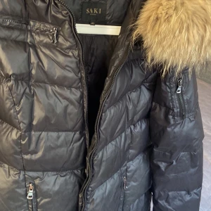 Svart dun jacka.st:40 m.päls Saki - Säljer en svart pufferjacka från Saki med snygg päls på huvan. Jackan har flera dragkedjefickor, quiltad design och är perfekt för kalla dagar. Stängs med dragkedja och har en modern, lite längre passform.passar storlek 38-40.1 gång använd.helt ny