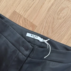 Svarta skinnbyxor från NA-KD med slits strl 36 - Snygga svarta byxor i skinnimitation från NA-KD. De har raka ben med slits nertill och en clean midja utan synliga fickor. Perfekta för dig som vill ha en edgy och trendig look.