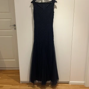 Marinblå långklänning med spetsdetaljer - Elegant marinblå långklänning med v-ringad hals och ärmlös design. Klänningen har vackra spetsdetaljer upptill och ett flowy tylltyg nedtill som ger en lyxig känsla. Perfekt för bal eller festliga tillfällen när du vill sticka ut.