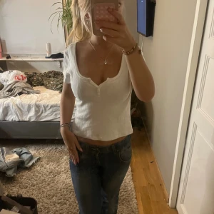 Vit ribbad topp med knappar - Säljer en söt vit ribbad topp från Brandy Melville med korta ärmar och liten v-ringning framtill med två knappar. Toppen är croppad och har diskret hålmönster i tyget. Perfekt till jeans eller kjol för en fräsch look.