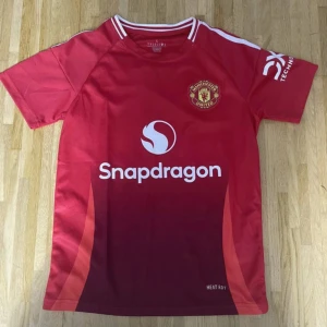 Manchester United röd fotbollströja S - Manchester Uniteds officiella matchtröja i rött från Adidas med Snapdragon-logga på bröstet och klubbmärke på vänster sida. Tröjan har vita detaljer vid krage och axlar samt korta ärmar. Tillverkad i lätt och ventilerande funktionsmaterial.