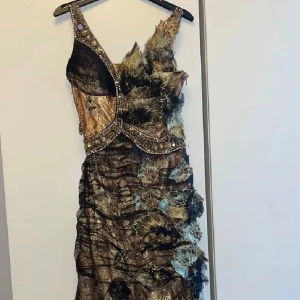 Unik långklänning med paljetter ( Balklänning)  - Säljer en extravagant långklänning i svart och guld med asymmetrisk design och stora blommiga detaljer. Klänningen har glittriga paljetter längs axelbanden och midjan, samt transparenta partier och texturerade lager. Perfekt för dig som vill sticka ut och älskar statement-plagg.