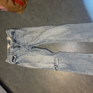 Ljusblå jeans från H&M med hål - Ljusblå jeans från H&M i klassisk femficksmodell med knäppning fram. Byxorna har ett snyggt slitet hål på ena benet och raka ben. Perfekta för en avslappnad och trendig look.