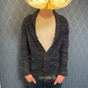 Mörkblå stickad kofta från Cotton Club - Säljer en mörkblå stickad kofta från Cotton Club med snyggt melerat mönster och knappar framtill. Koftan har långa ärmar med mörka armbågslappar och bred krage. Perfekt för lager-på-lager och chill stil. Tröjan har ett litet hål men det är syns knappt när man har den på sig. Om du har några frågor är det bara att höra av sig, inga prisförslag är dåliga! 