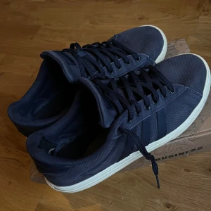 Mörkblå sneakers från Adidas - Snygga mörkblå sneakers från Adidas med klassiska tre ränder på sidan. Ovandel i textil och snörning framtill. Skorna har vit platt sula och en stilren, enkel design som passar till det mesta. Perfekta för dig som gillar en clean och sportig look. Och är lite trasig inuti som i bilden