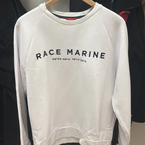 Vit sweatshirt från Race Marine L - Vit sweatshirt från Race Marine med svart tryck framtill och koordinater i rött och svart. Tröjan har rund halsringning, långa ärmar och mjuk bomullskänsla. Perfekt för en clean och sportig stil.