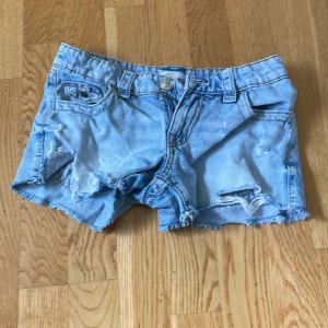 Ljusblå jeansshorts från Gina Tricot - Säljer ett par ljusblå jeansshorts från Gina Tricot med slitna detaljer och råa kanter. Shortsen har snygga broderier på bakfickorna. Tyvärr har det spruckit lite på vänster sida.